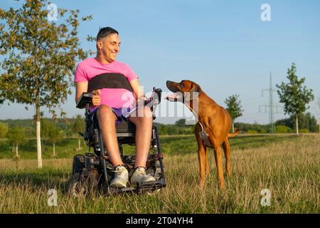 Uomo in sedia a rotelle che parla con un adorabile compagno di cani seduto sull'erba. Proprietario di sesso maschile che si gode un weekend con il cane sullo sfondo soleggiato della campagna Foto Stock