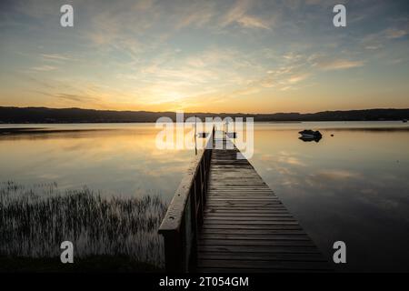 Un molo di legno si estende graziosamente sopra le calme acque della laguna, rispecchiando nuvole dipinte all'alba, con montagne lontane all'orizzonte Foto Stock