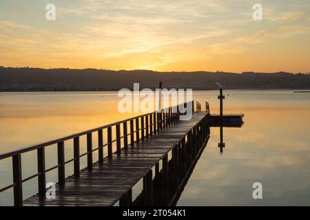 Un molo di legno si estende graziosamente sopra le calme acque della laguna, rispecchiando nuvole dipinte all'alba, con montagne lontane all'orizzonte Foto Stock