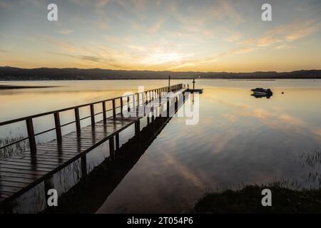 Un molo di legno si estende graziosamente sopra le calme acque della laguna, rispecchiando nuvole dipinte all'alba, con montagne lontane all'orizzonte Foto Stock