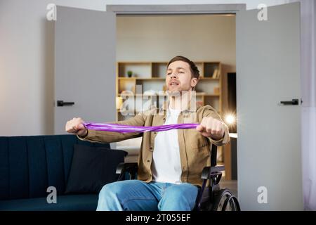 Giovane disabile in sedia a rotelle facendo esercizi con elastico a casa. Ragazzo caucasico handicappato che lavora in soggiorno. Foto Stock