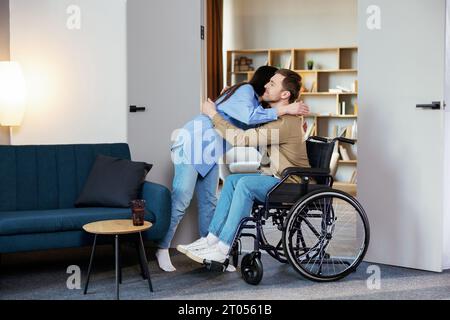 Giovane uomo in sedia a rotelle e sua moglie che si abbraccia, in salotto a casa. Foto Stock
