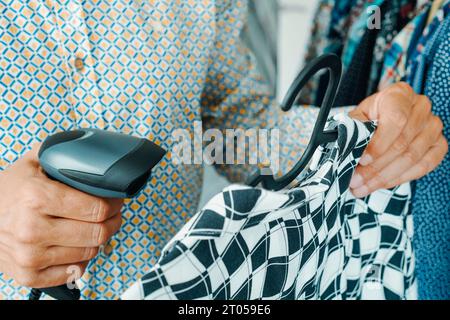 primo piano di un assistente del negozio, in un negozio di abbigliamento, scansionando il codice a barre di una camicia bianca e nera appesa a un appendiabiti utilizzando un lettore di codici a barre Foto Stock