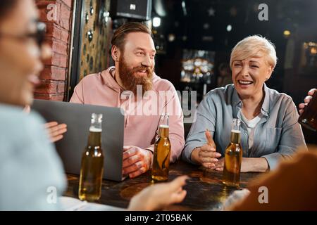 uomo barbuto e donna di mezza età che ridono vicino al portatile e donna afroamericana che parla in un pub Foto Stock