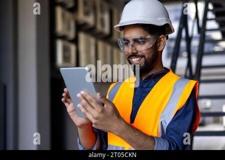 Ritratto di un giovane ingegnere, uomo in casco e giubbotto utilizzando un tablet, sorridendo, controllando i dati di produzione. Foto Stock