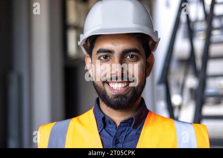 Ritratto ravvicinato di un giovane ingegnere in elmetto e giubbotto, uomo sorridente e guardante la macchina fotografica, operaio in fabbrica e magazzino logistico. Foto Stock