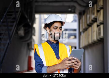 Ritratto di un giovane ingegnere, uomo in casco e giubbotto utilizzando un tablet, sorridendo, controllando i dati di produzione. Foto Stock