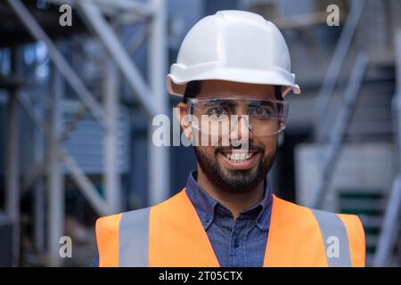 Ritratto ravvicinato di un giovane ingegnere in elmetto e giubbotto, uomo sorridente e guardante la macchina fotografica, operaio in fabbrica e magazzino logistico. Foto Stock