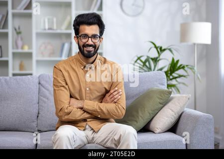 Ritratto di un giovane indiano sorridente seduto a casa sul divano con le braccia incrociate e guardando con sicurezza la fotocamera. Foto Stock