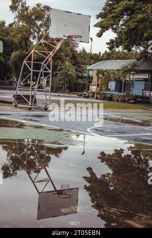 Il tabellone da basket in legno con il cerchio in metallo e la rete bianca mostrano il loro riflesso sull'acqua piovana su tutto il vecchio cemento da basket Foto Stock