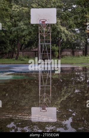 Il tabellone da basket in legno con il cerchio in metallo e la rete bianca mostrano il loro riflesso sull'acqua piovana su tutto il vecchio cemento da basket Foto Stock