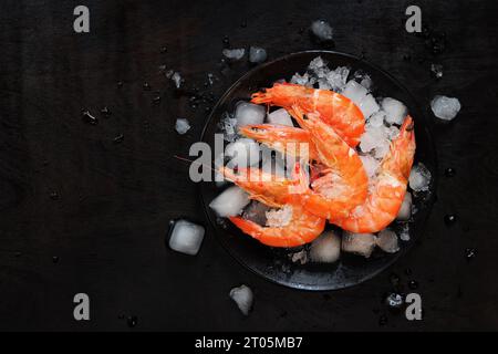 Grandi gamberi freddi in un piatto con ghiaccio su fondo di legno scuro. Gamberi bolliti in un piatto nero con ghiaccio. Gamberi freschi su sfondo nero, top Foto Stock