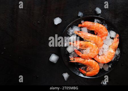 Grandi gamberi freddi in un piatto con ghiaccio su fondo di legno scuro. Gamberi bolliti in un piatto nero con ghiaccio. Gamberi freschi su sfondo nero, top Foto Stock