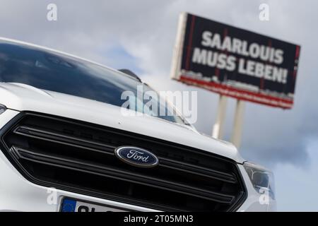 Saarlouis, Germania. 4 ottobre 2023. Una Ford Kuga si trova di fronte a un grande cartello che pubblicizza l'importanza dello stabilimento Ford di Saarlouis. Durante una riunione di lavoro, la forza lavoro spera di saperne di più sul futuro dello stabilimento. Crediti: Oliver Dietze/dpa/Alamy Live News Foto Stock