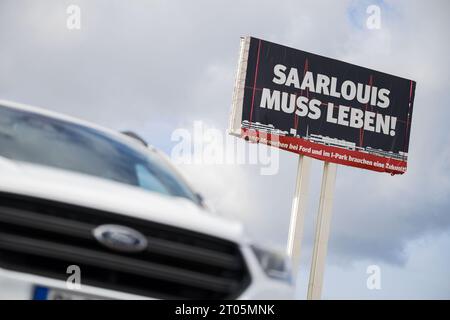 Saarlouis, Germania. 4 ottobre 2023. Una Ford Kuga si trova di fronte a un grande cartello che pubblicizza l'importanza dello stabilimento Ford di Saarlouis. Durante una riunione di lavoro, la forza lavoro spera di saperne di più sul futuro dello stabilimento. Crediti: Oliver Dietze/dpa/Alamy Live News Foto Stock