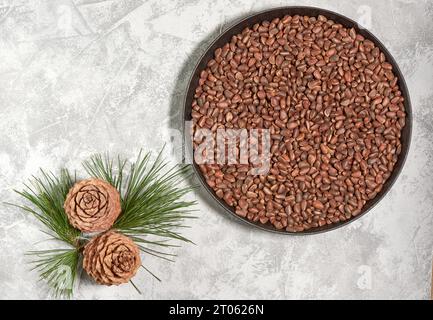 Noci di cedro o pino in una ciotola con coni su sfondo grigio e un ramo di un albero di cedro con aghi verdi, vista dall'alto Foto Stock