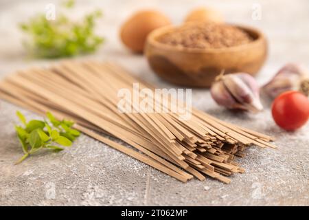 Tagliatelle giapponesi di soba di grano saraceno con pomodoro, uova, spezie, erbe su fondo marrone di cemento. Vista laterale, primo piano, messa a fuoco selettiva. Foto Stock