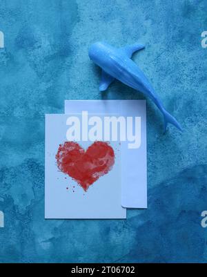 una natura morta con una carta con un cuore acquerello incorniciato da balene giocattolo su uno sfondo blu acquerello Foto Stock