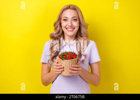 Ritratto di donna ottimista con capelli ondulati e t-shirt viola con cesto di fragole isolato su sfondo giallo Foto Stock