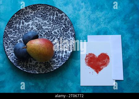 una natura morta con un piatto con motivo floreale, pera matura, prugne e un cartoncino di carta con cuore ad acquerello su sfondo blu Foto Stock