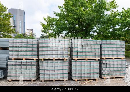 Lastre di pavimentazione in calcestruzzo impilate su pallet in legno in un cantiere edile. Alberi ed edifici sullo sfondo. Foto Stock