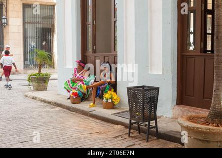 Due donne di intrattenimento in abiti tradizionali cubani si siedono a due passi dall'Avana Vecchia. Hanno cesti con fiori. Foto Stock