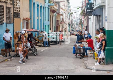 Un cubano vende prodotti alimentari con un piccolo carrello in un angolo della città. Lo stile di vita urbano di altre persone è mostrato nella scena della strada. Foto Stock