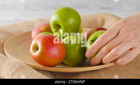 Mele rosse e verdi su un piatto di legno da vicino. Mani femminili che tengono una mela Foto Stock