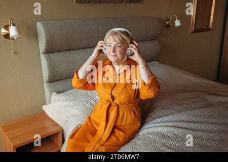 Una donna anziana positiva siede sul letto, ascolta la musica con le cuffie wireless e balla. Foto Stock