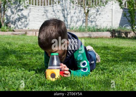 il bambino in età prescolare sdraiato sullo stomaco sul prato verde esamina un insetto utilizzando un pallone giocattolo educativo con occhiali da ingrandimento. Il bambino esplora il mondo Foto Stock