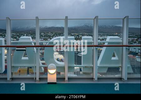 Logo AIDA attaccato alla ringhiera esterna della nave da crociera AIDA bella, città dell'Islanda sullo sfondo, in serata Foto Stock