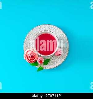 Layout creativo composto da una tazza di tè decorata con fiori rosa e foglie verdi su sfondo blu chiaro. Concetto minimale di bevanda calda e fiori Foto Stock