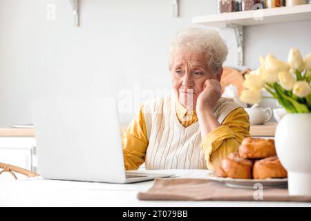 Sconvolta una donna anziana che usa un computer portatile al tavolo in cucina Foto Stock