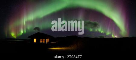 La luminosa e colorata aurora boreale, l'aurora boreale, balla attraverso un cielo aperto con una cabina in primo piano e luci accese nelle finestre Foto Stock