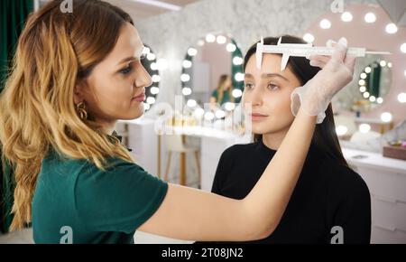 Estetista che utilizza lo strumento di misurazione per la procedura di correzione sopracciglia nel salone di bellezza. Specialista di bellezza in guanti sterili che posizionano il righello sulla fronte della donna durante le misurazioni per la correzione delle sopracciglia. Foto Stock