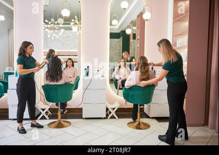 Due parrucchieri femmina arricciano i capelli del cliente e fanno l'acconciatura nel salone di bellezza moderno. Giovani donne sedute in sedie di fronte agli specchi mentre parrucchieri styling capelli dei clienti. Foto Stock