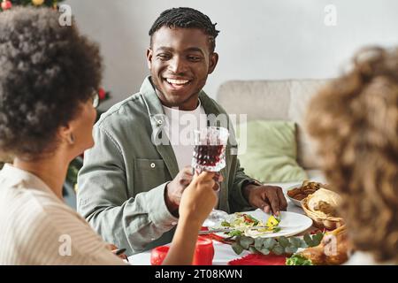 Allegro uomo afroamericano seduto a un tavolo festivo circondato dai suoi parenti, Natale Foto Stock