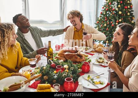 La famiglia allegra e multirazziale ride e sorride durante il pranzo festivo con l'albero di Natale sullo sfondo Foto Stock