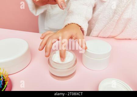 Foto ritagliata da vicino della mano di una bambina, un dito in un barattolo di panna, lozione su sfondi rosa. Bambino imparare prendersi cura di bellezza, salute. Foto Stock