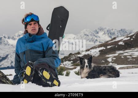 Giovane uomo con abbigliamento sportivo e snowboard seduto su un terreno innevato vicino a cani bianchi e neri mentre guarda lontano dalle montagne nelle nuvolose giornate invernali Foto Stock