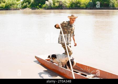 Pescatore concentrato con un simpatico cane che si allontana dal molo con un lungo bastone, mentre naviga per pescare. Foto Stock