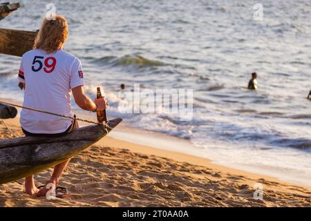 Il turista sta sta beve una birra Lion seduto su una barca di pescatori sulla spiaggia di Negombo in Sri Lanka al tramonto Foto Stock