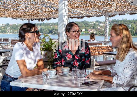 Tre amiche allegri siedono a un tavolo per una pausa caffè, chiacchierano e ridono sulla terrazza di un ristorante sul fiume. Foto Stock
