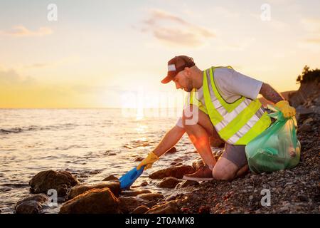 Vista laterale di un uomo caucasico barbuto adulto volontario che indossa giubbotto e guanti di gomma per prelevare la bottiglia di plastica dal mare. Concetto di giornata della Terra e en Foto Stock