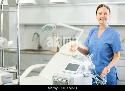 Ritratto di mezza età sorridente massaggiatrice medico donna in piedi vicino alla macchina di bellezza in salone medico, stretching mani da parte con il palmo aperto, Foto Stock