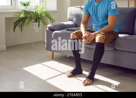 Uomo seduto sul divano in calze di compressione medica sulle gambe per la prevenzione delle vene varicose. Foto Stock