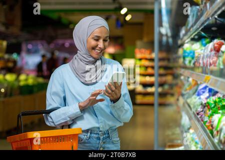 La giovane donna felice che acquista con il telefono sceglie i prodotti in un grande negozio di alimentari, la donna musulmana in hijab utilizza la lista della spesa online e utilizza l'app sullo smartphone. Foto Stock