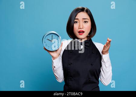 Cameriera asiatica che tiene sveglia con un ritratto sorpreso dell'espressione facciale. Preoccupata giovane dipendente del caffè che indossa un'uniforme professionale che mostra il tempo e guarda la macchina fotografica Foto Stock