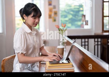 L'immagine laterale di una giovane donna asiatica che lavora in remoto in una bella caffetteria si concentra sul suo lavoro sul computer portatile. Lifestyle co Foto Stock