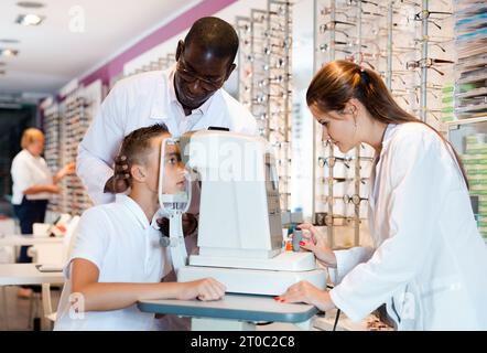 Due optometristi qualificati che utilizzano il rifrattometro per testare la vista di ragazzo teen in negozio di ottica Foto Stock